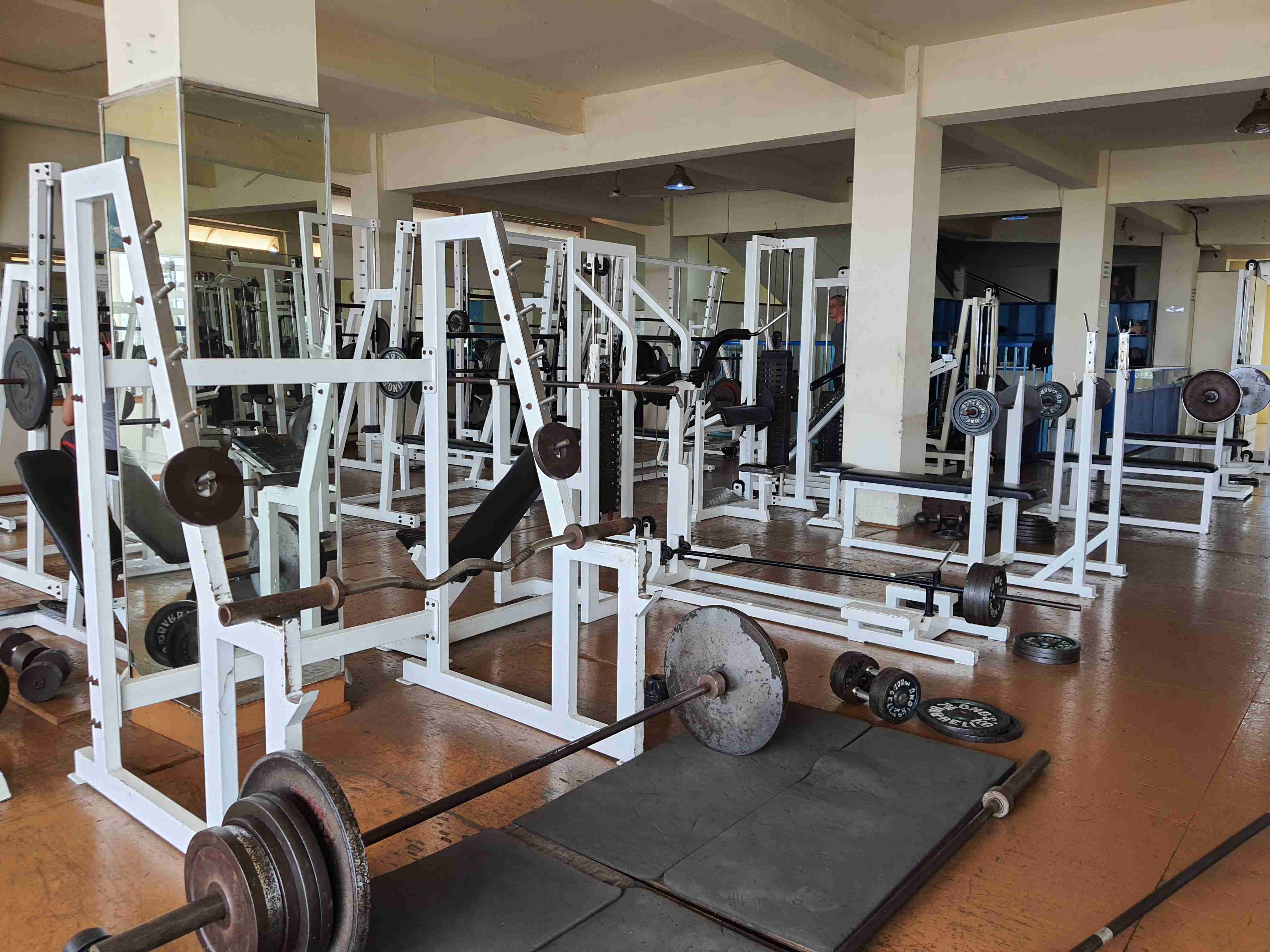 Foto sebagian alat fitness di Tetap Segar Fitness dengan highlight di squat rack dan banyak pilihan bar.